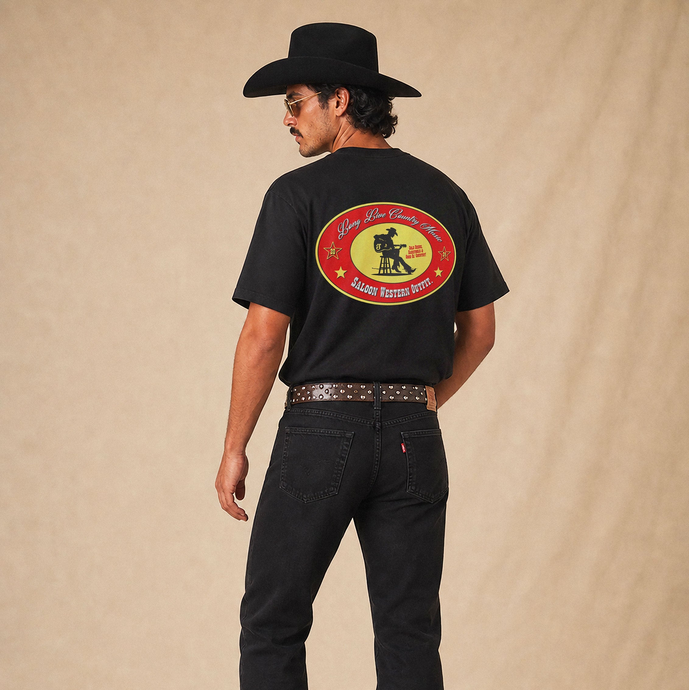 A man wearing a black cowboy hat, black boots, and black jeans stands with his back to the camera, wearing a black t-shirt with a large red, yellow, and white circular graphic on the back that reads 'Long Live Country Music' in an emblem branded with Saloon.